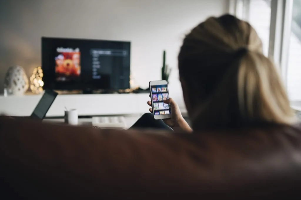 Person browsing a streaming app on a phone while relaxing on a couch in front of a TV